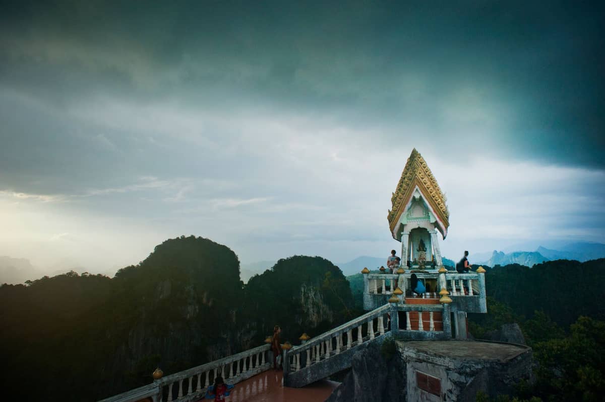 Tempelgipfel und Besucher auf dem Tiger Cave Temple mit dramatischer Berglandschaft und bewölktem Himmel in Krabi, Thailand.
