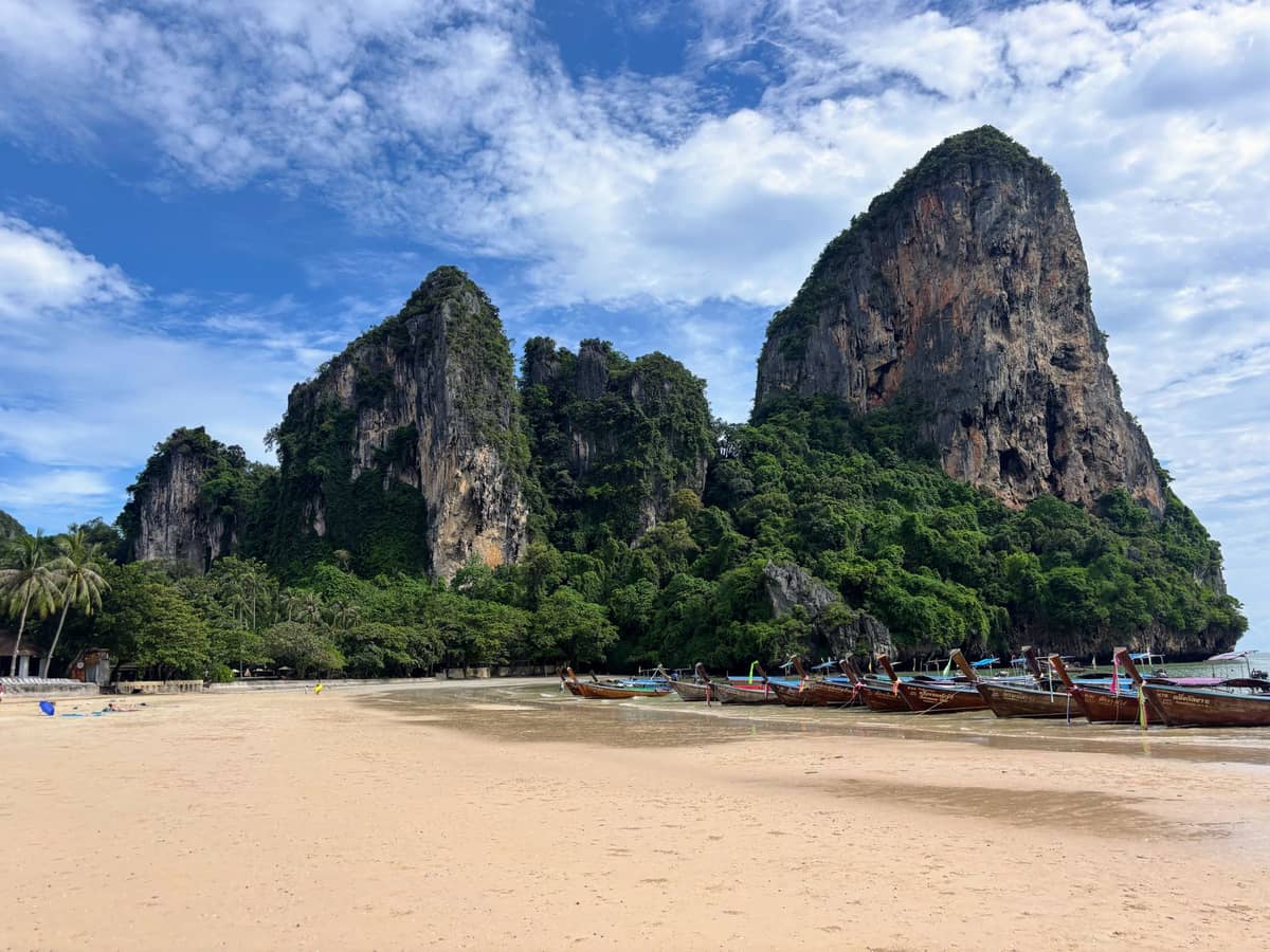 Breiter Sandstrand in Krabi mit markanten Kalksteinfelsen und Longtail-Booten am Ufer unter einem teils bewölkten blauen Himmel.