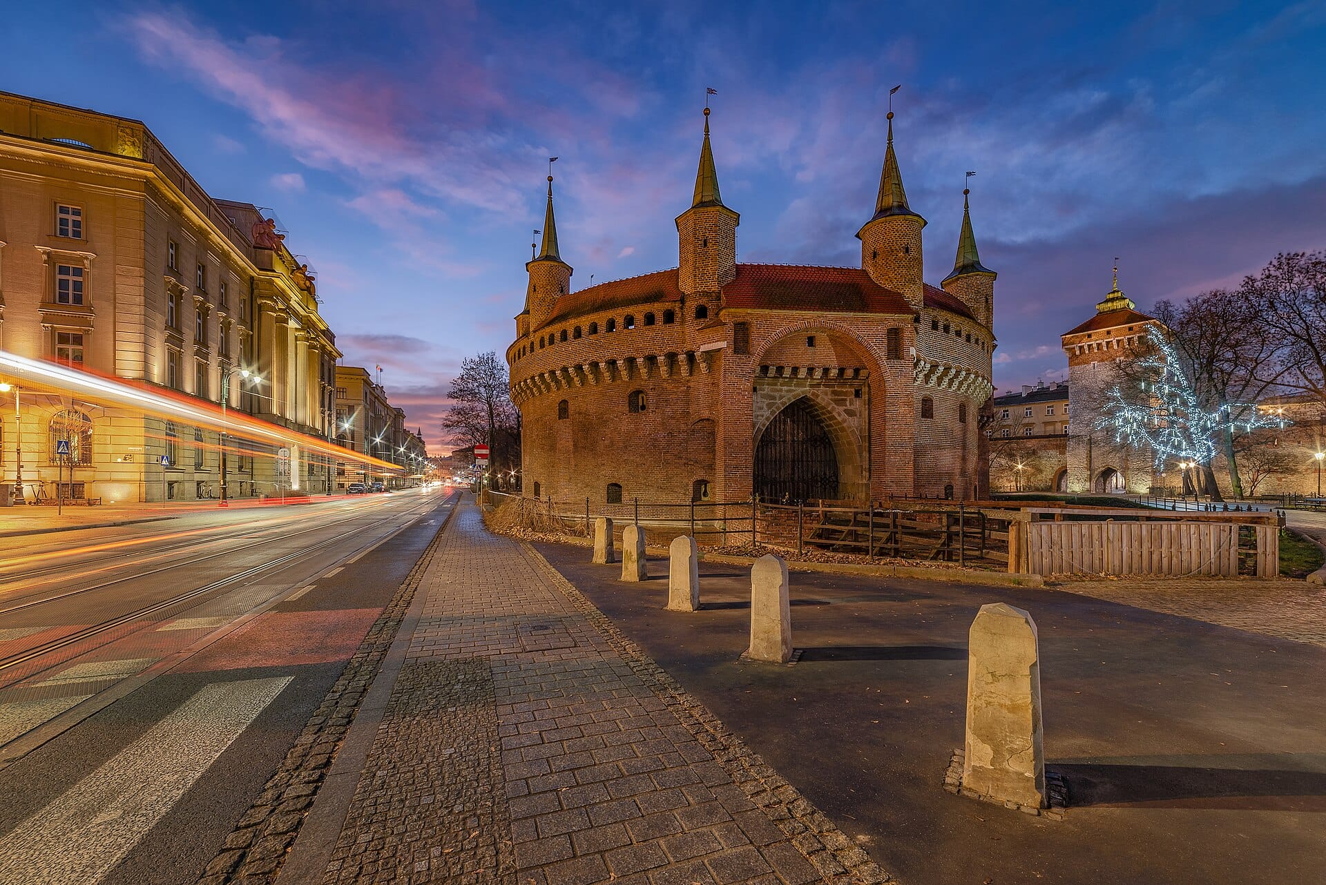วิว Barbakan คราคูฟช่วงพลบค่ำ เห็นผนังอิฐสีแดง ทางเข้าโค้ง และยอดป้อมปลายแหลมสีเขียวในฉากเมือง