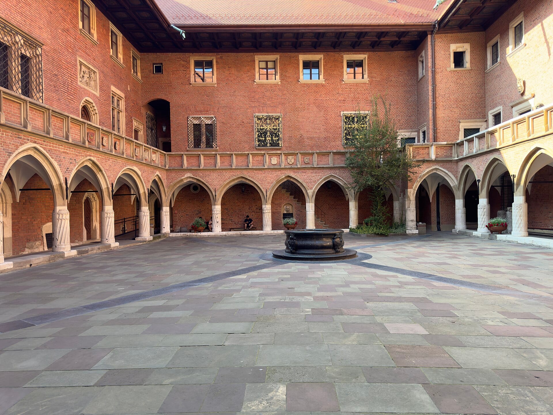 ลานกลางแจ้งสไตล์ยุคกลางอันสงบงามของ Collegium Maius ที่มีซุ้มโค้งหิน กำแพงอิฐแดง และบ่อน้ำกลางลานใน เมืองเก่าคราคูฟ