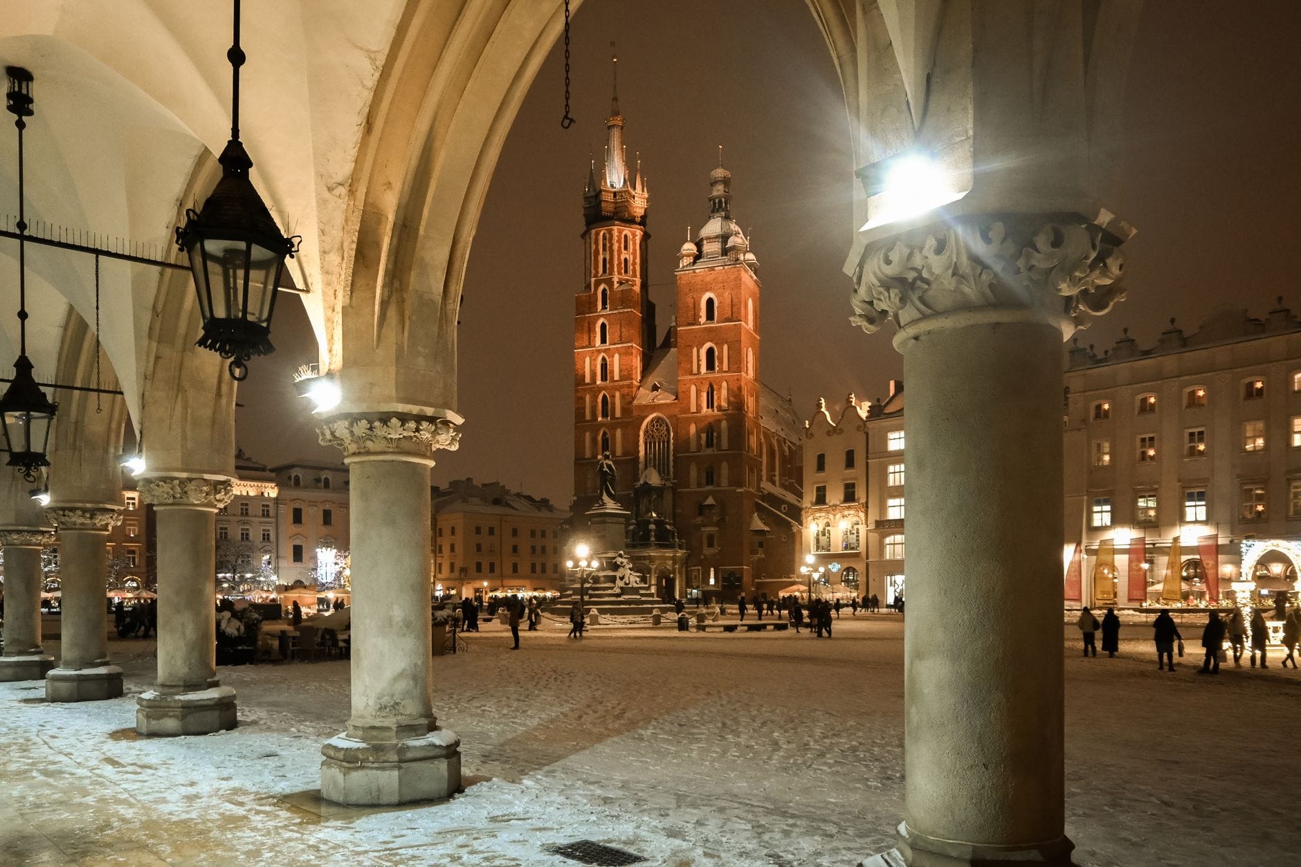 วิวยามค่ำคืนของจัตุรัส Rynek Główny ในเมือง Kraków พร้อมมหาวิหาร มหาวิหารเซนต์แมรี่ ที่สว่างไสว มองผ่านซุ้มโค้งของ Sukiennice และผู้คนเดินเล่นในหิมะ
