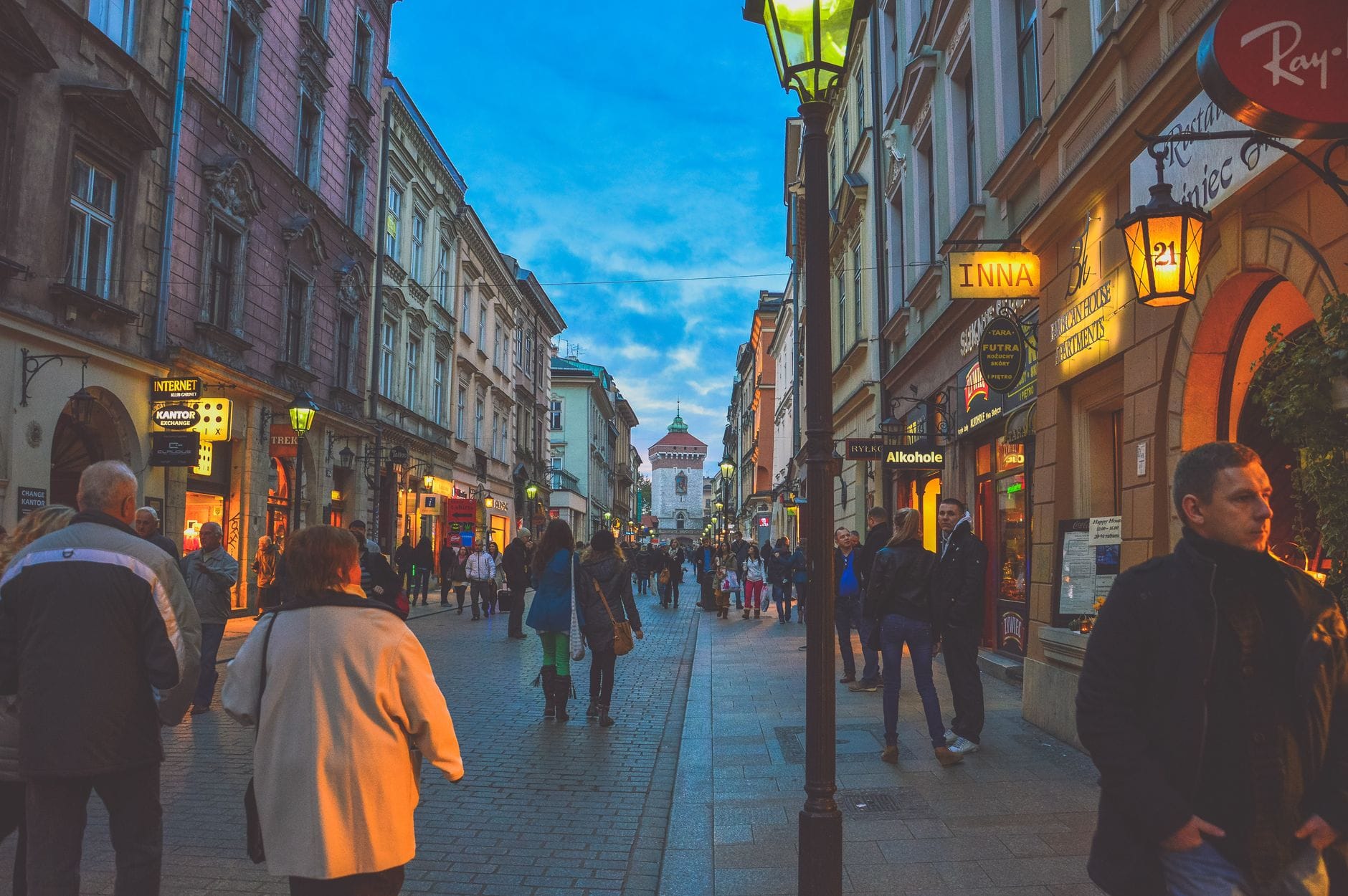 ผู้คนเดินเล่นบนถนนที่คึกคักของเมือง Kraków ซึ่งเรียงรายด้วยบาร์ ร้านค้า และแสงไฟยามเย็นที่สดใส สื่อถึงบรรยากาศการสังสรรค์ยามค่ำคืน
