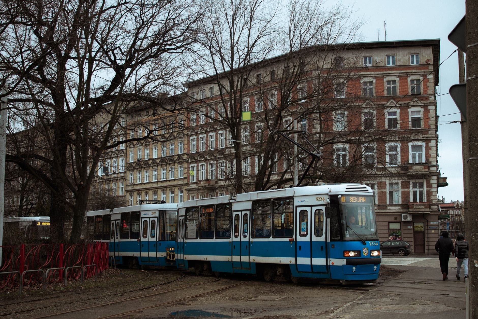 รถรางสีน้ำเงินและขาววิ่งผ่านย่านที่พักอาศัยในเมือง Kraków พร้อมอาคารอพาร์ตเมนต์แบบคลาสสิกและต้นไม้ผลัดใบในฤดูหนาว