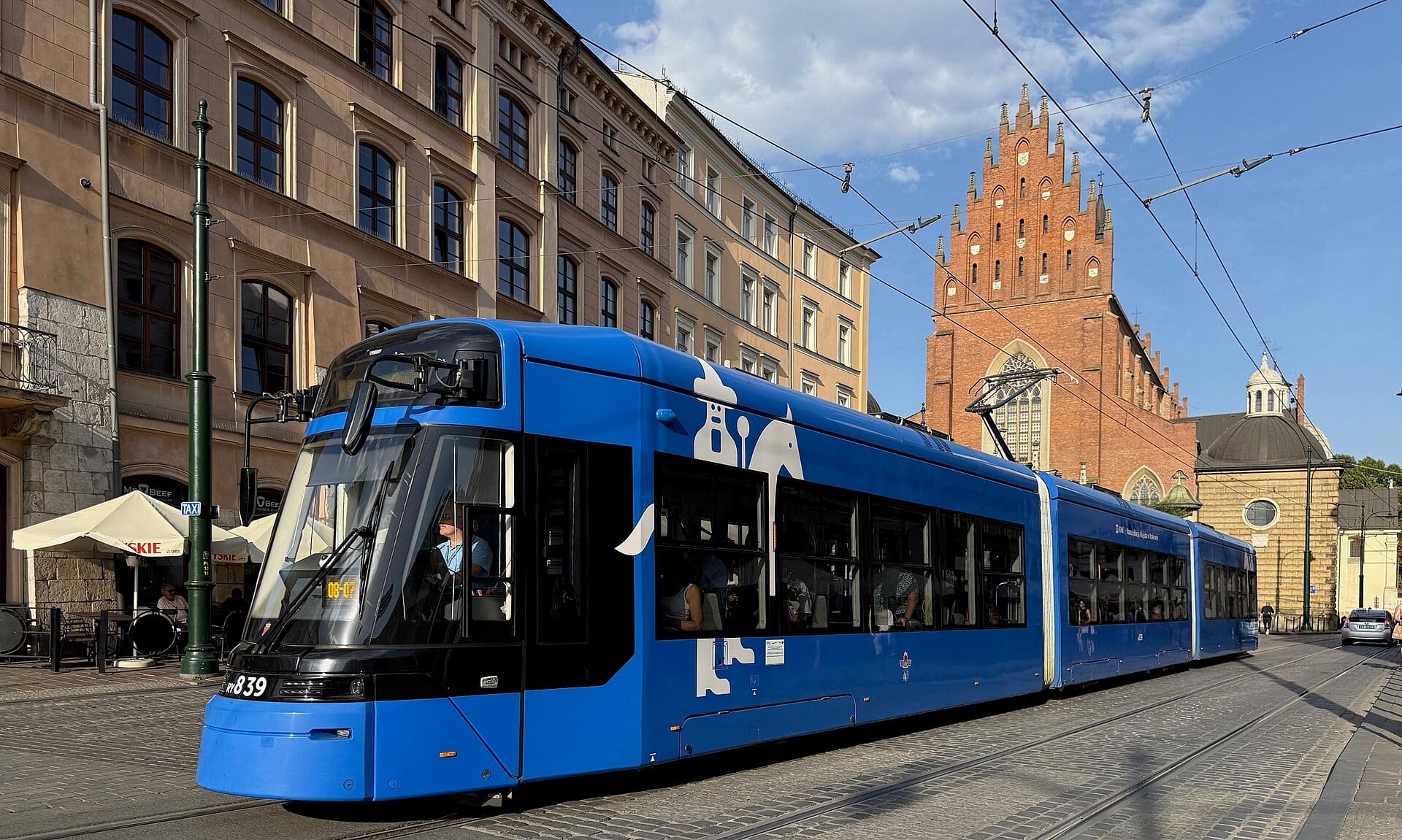 รถรางสีน้ำเงินสมัยใหม่วิ่งผ่านใจกลางเมือง Kraków พร้อมโบสถ์อิฐประวัติศาสตร์และอาคารในเมืองเป็นฉากหลังในวันที่แดดจัด