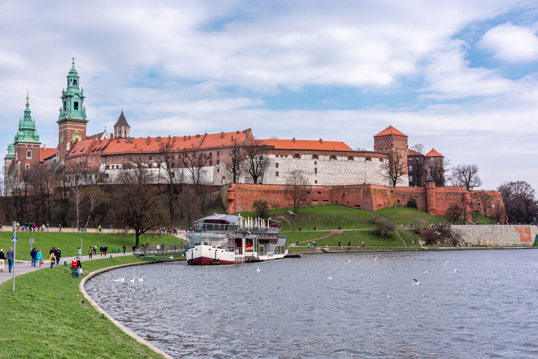 ปราสาท Wawel ในเมือง Kraków มองจากฝั่งตรงข้ามของแม่น้ำ Vistula พร้อมเส้นทางเดินเท้า ผู้คน และเรือนำเที่ยวบริเวณริมน้ำ