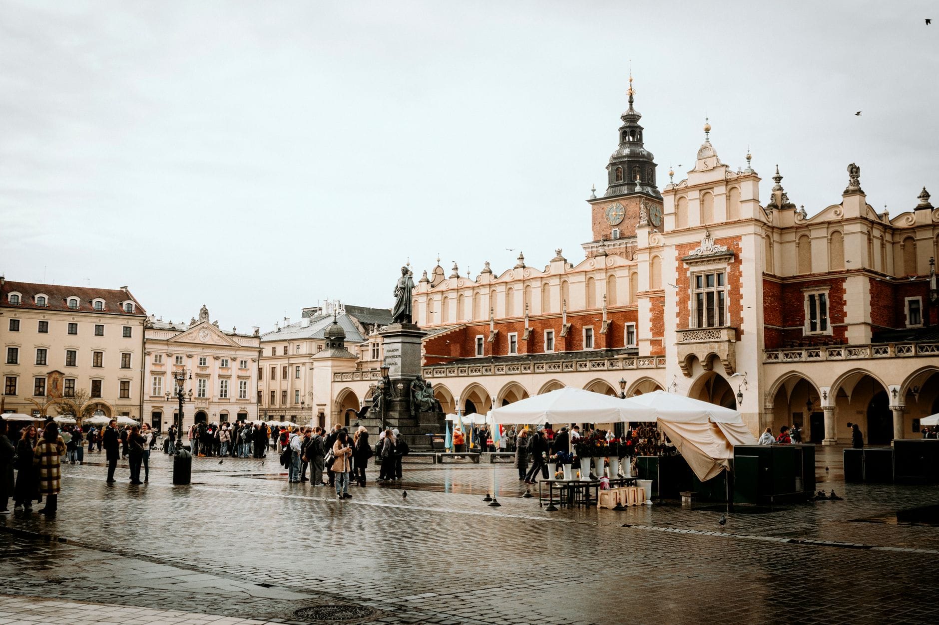 มุมมองกว้างแนวนอนของ Rynek Główny ในเมือง Kraków พร้อมอาคาร Cloth Hall ที่มีประวัติศาสตร์และแผงตลาด คึกคักไปด้วยผู้คนในวันที่มีเมฆมาก