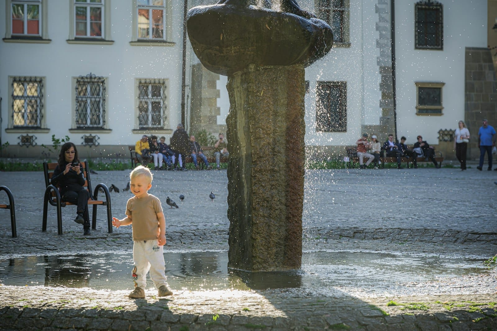 เด็กเล็กเล่นอยู่ใกล้น้ำพุหินในจัตุรัสที่แดดสดใสของ Kraków โดยมีอาคารประวัติศาสตร์และผู้คนที่นั่งพักผ่อนบนม้านั่งเป็นฉากหลัง