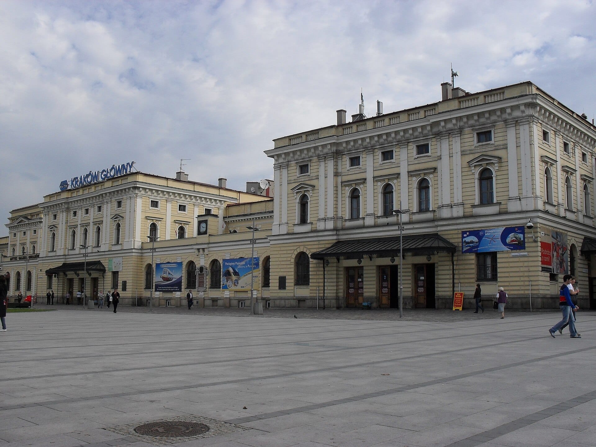 ด้านหน้าของสถานีรถไฟหลัก Kraków Główny อาคารประวัติศาสตร์สีเหลืองอ่อนพร้อมผู้คนเดินอยู่ด้านหน้า