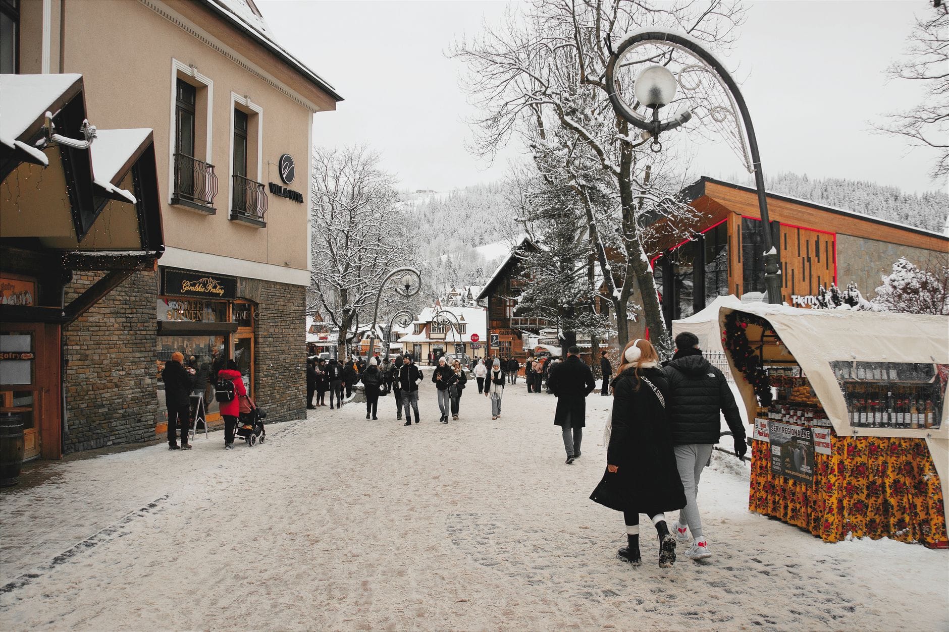 ถนนคนเดิน Krupówki ในเมือง Zakopane มีร้านค้าและแผงอาหารเรียงราย ผู้คนเดินในวันที่หิมะตก โดยมีภูเขาเป็นฉากหลัง