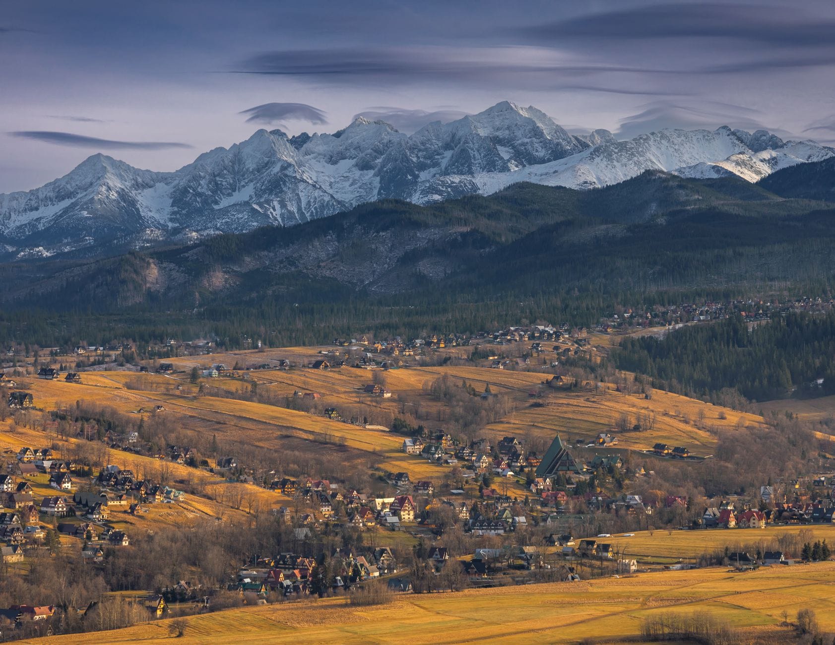 วิวกว้างของเมือง Zakopane ท่ามกลางทุ่งสีทองในฤดูใบไม้ร่วง พร้อมเทือกเขา Tatra ที่ปกคลุมด้วยหิมะในฉากหลังใต้ท้องฟ้าที่มืดครึ้ม
