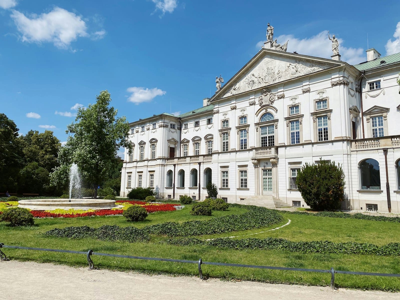 พระราชวัง Krasiński ในกรุง Warsaw พร้อมด้วยด้านหน้าอาคารสไตล์บาโรกสีขาว สวนสีเขียวขจี แปลงดอกไม้หลากสีสัน และน้ำพุกลางลานภายใต้ท้องฟ้าสีฟ้าสดใส