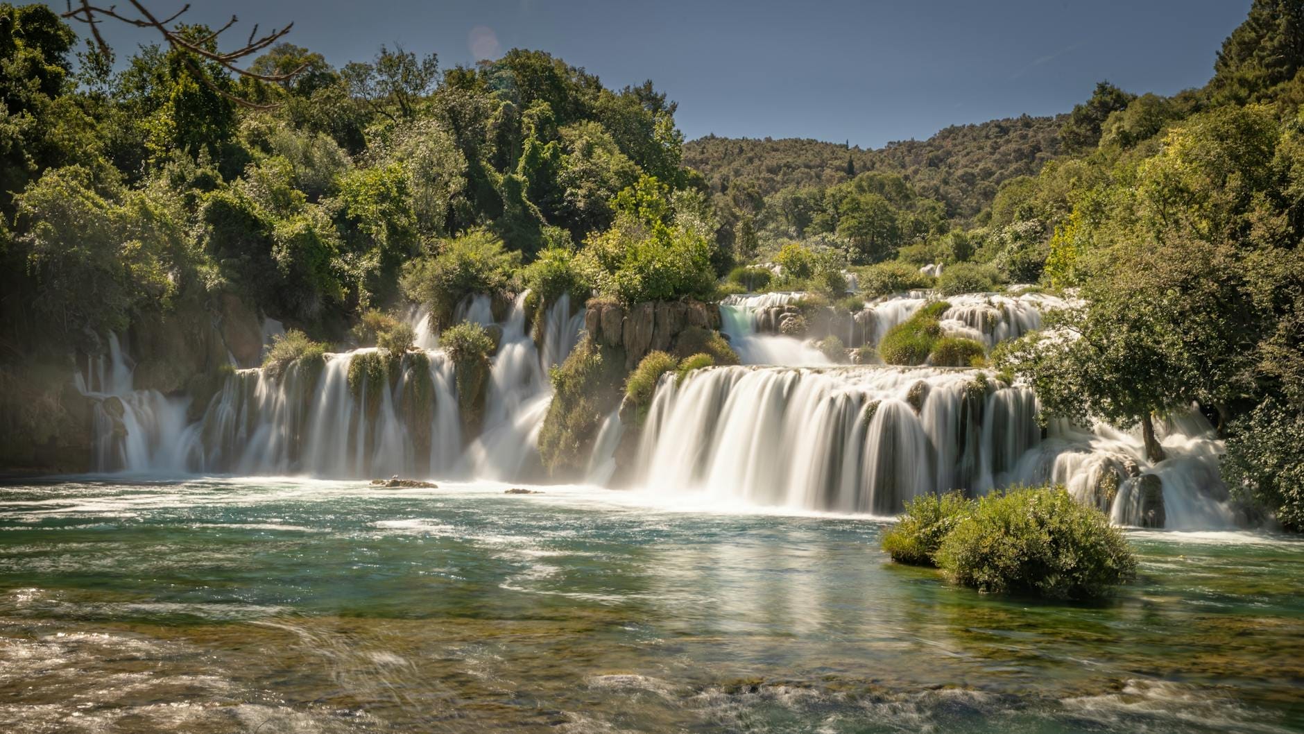 Vista panorámica de las cascadas de Kravica cayendo sobre acantilados de travertino hacia una piscina turquesa rodeada de frondosa vegetación bajo un cielo despejado.