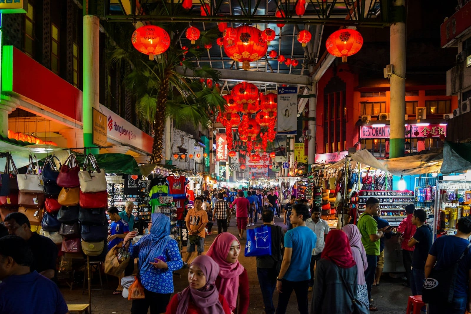 Shoppers browse vibrant street stalls under red lanterns at Kuala Lumpur’s bustling night market, with colorful bags and clothes displayed on either side.