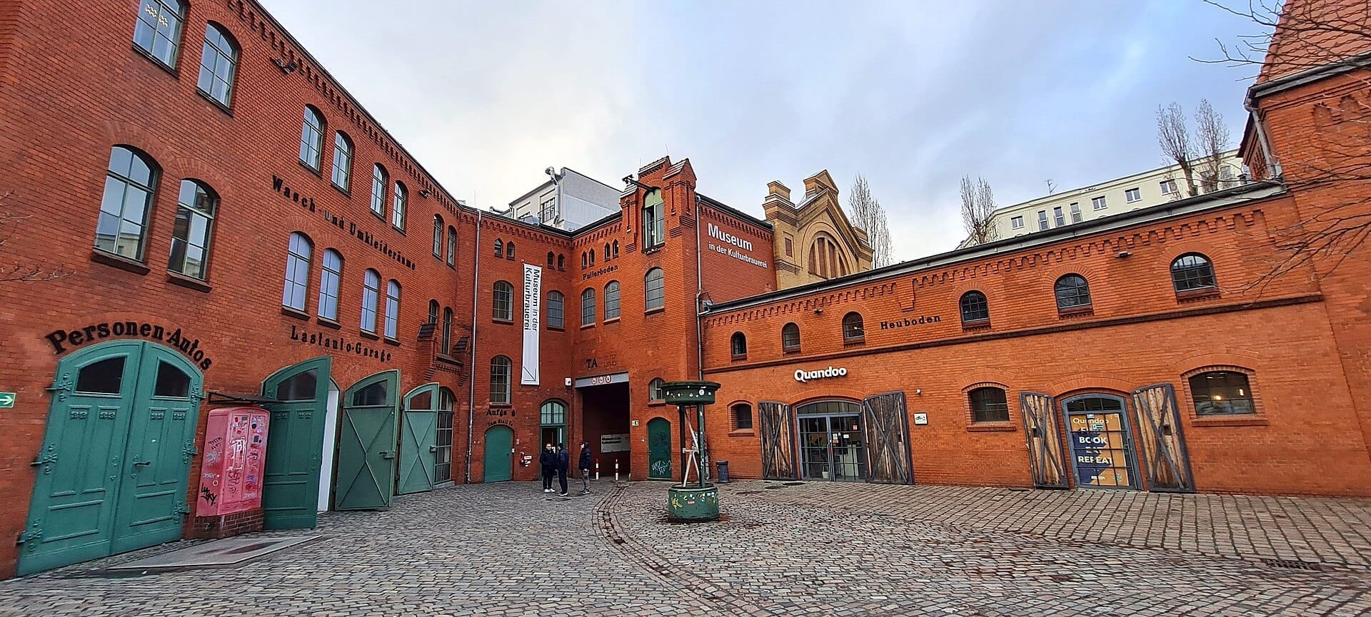 Vista degli edifici in mattoni rossi e del cortile acciottolato della Kulturbrauerei a Berlino, con porte verdi e cartelli del museo visibili.