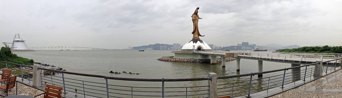 Panoramablick auf die Kun Iam Statue auf ihrem lotusförmigen Sockel, umgeben von Wasser und der Skyline der Stadt unter einem bewölkten Himmel in Macau.