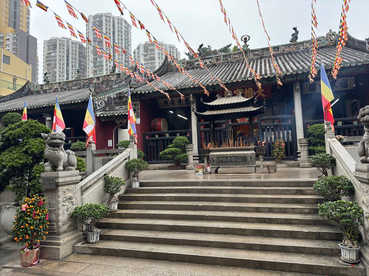 Eingangstreppe und Eingang des Kun Iam Tempels in Macau mit bunten Fahnen, Räuchergefäß, steinernen Löwenstatuen und traditionellen chinesischen Architekturdetails.