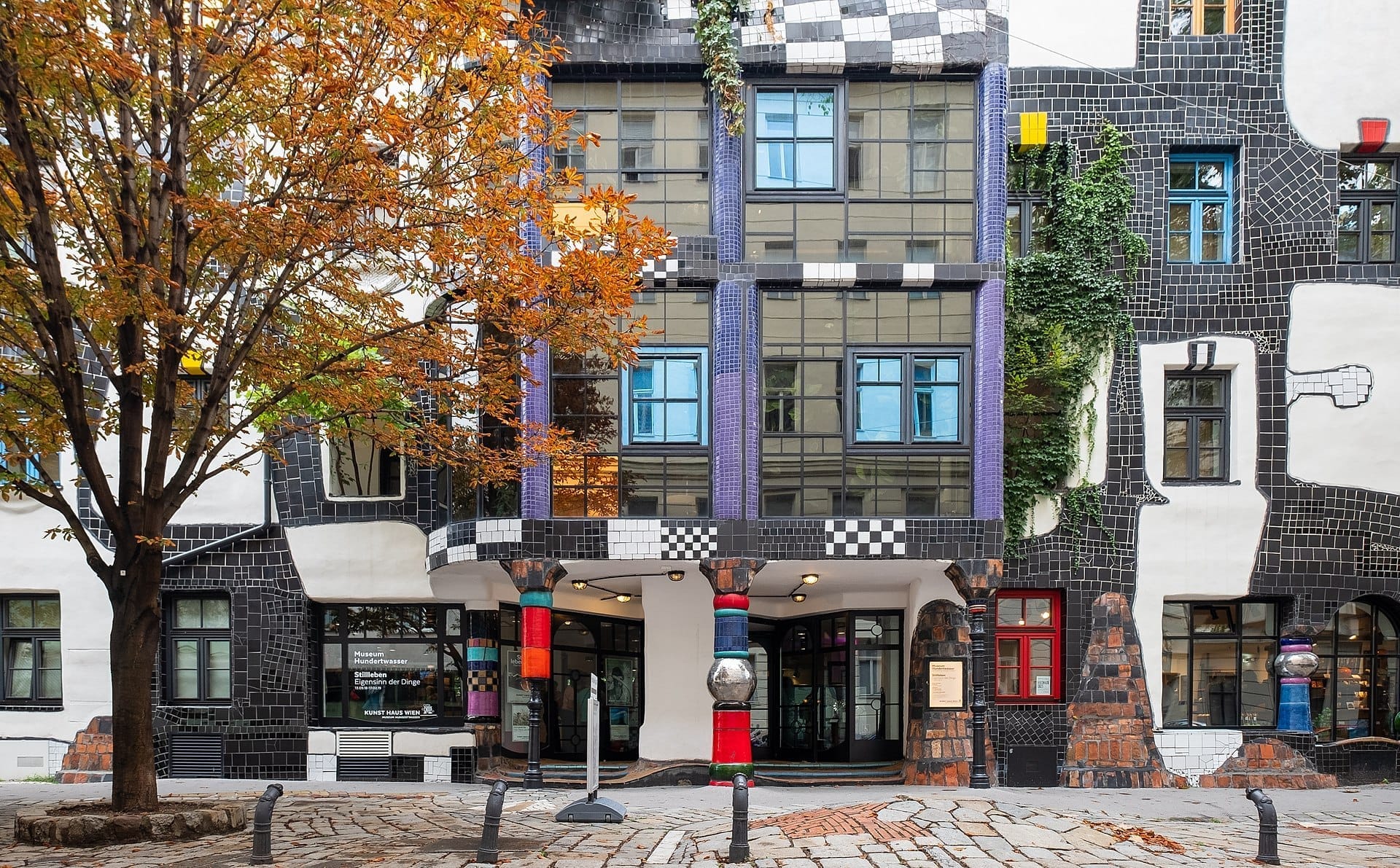 Frontansicht des Kunst Haus Wien mit bunten Säulen, Mosaikfassade und einem Herbstbaum, der den Museumseingang in Wien einrahmt.