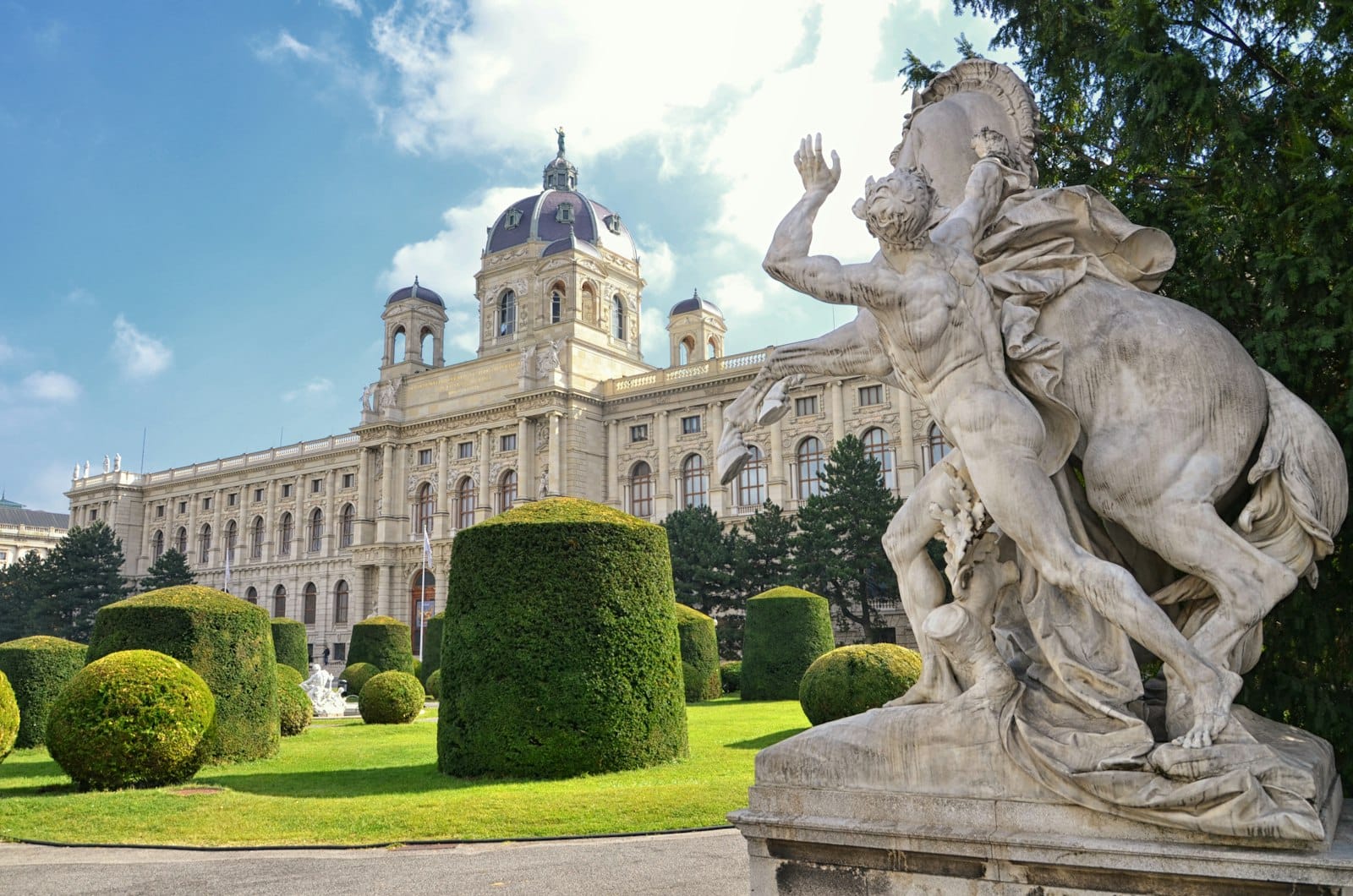 Außenansicht des Kunsthistorischen Museums Wien mit beeindruckender Neorenaissance-Kuppel, gepflegten Gartenanlagen und einer klassischen Marmorstatue vor blauem Himmel.