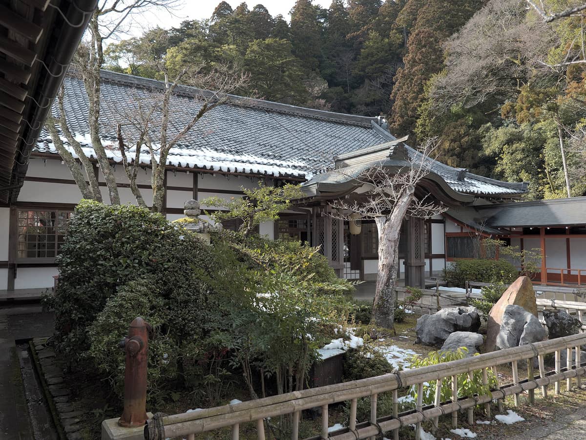 Traditional main building of Kurama-dera Temple surrounded by winter greenery and forested mountain slopes, snow patches visible in the peaceful courtyard.