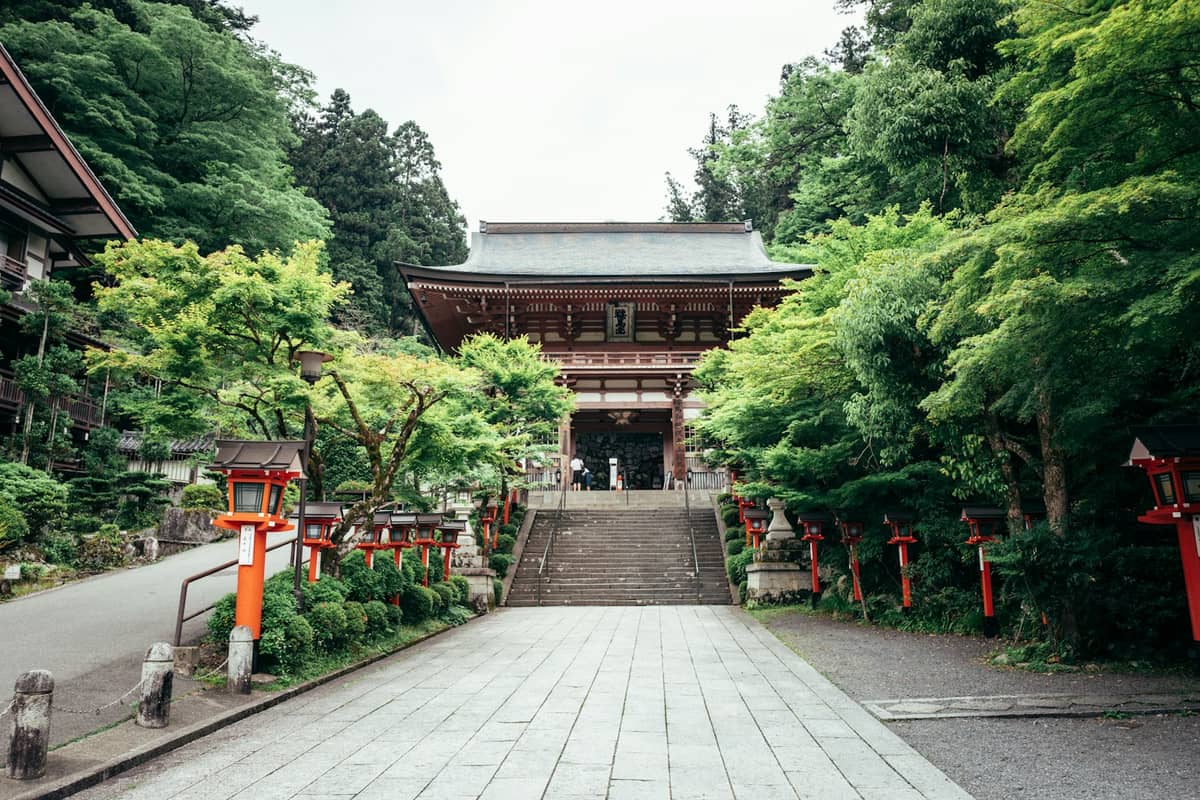 The main entrance and stone steps of Kurama-dera Temple, surrounded by lush green trees and red lanterns in Kurama village.