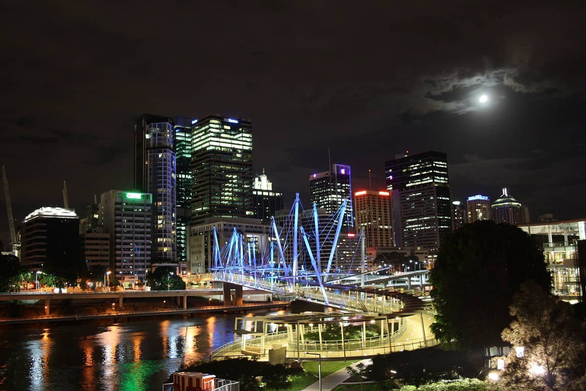 Die Kurilpa Bridge nachts beleuchtet, die den Brisbane River überspannt, mit hell erleuchteten Wolkenkratzern der Stadt und Spiegelungen auf dem Wasser unter einem bewölkten Himmel.
