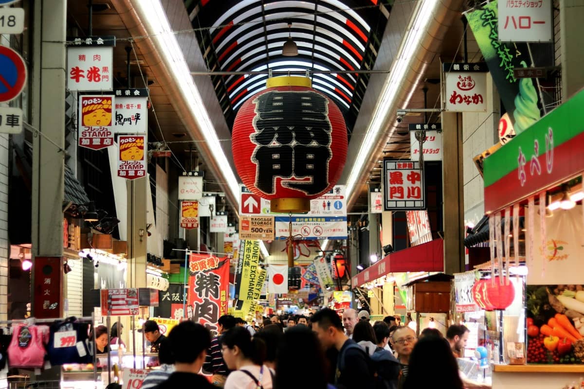 Crowds walk under colorful lights and a giant lantern inside Kuromon Ichiba Market, surrounded by food stalls and Japanese signs in Osaka.