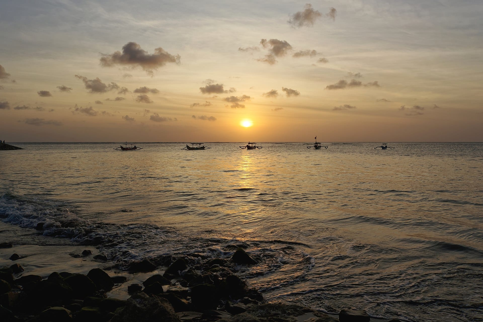 Vista panoramica del tramonto da Kuta Beach a Bali, con onde dolci e barche lontane all'orizzonte.