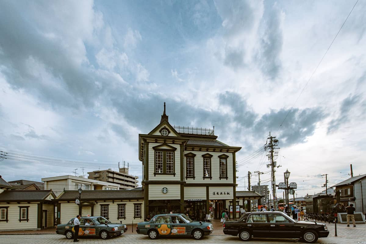 Several Japanese taxis waiting outside a historic-looking Kyoto train station under a cloudy sky.