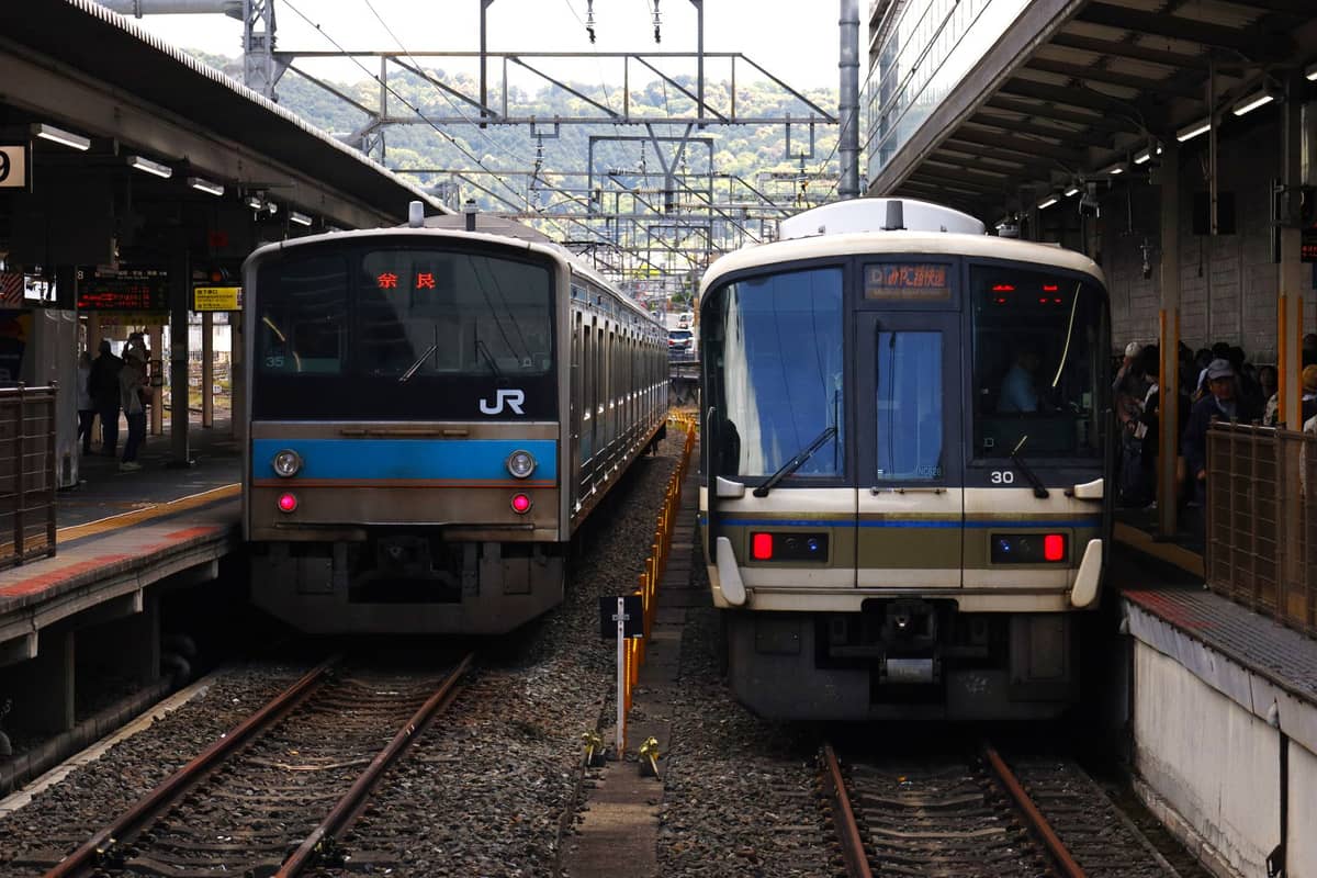 Two JR commuter trains at a platform in a Japanese rail station, passengers visible waiting for boarding. Overhead wires and mountain scenery in background.