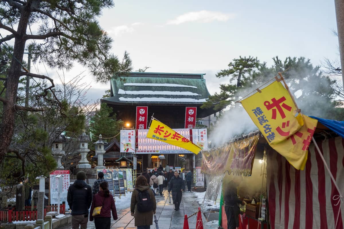 Food stalls with steam and yellow banners line a path towards a traditional Japanese shrine, with people walking and winter trees around.