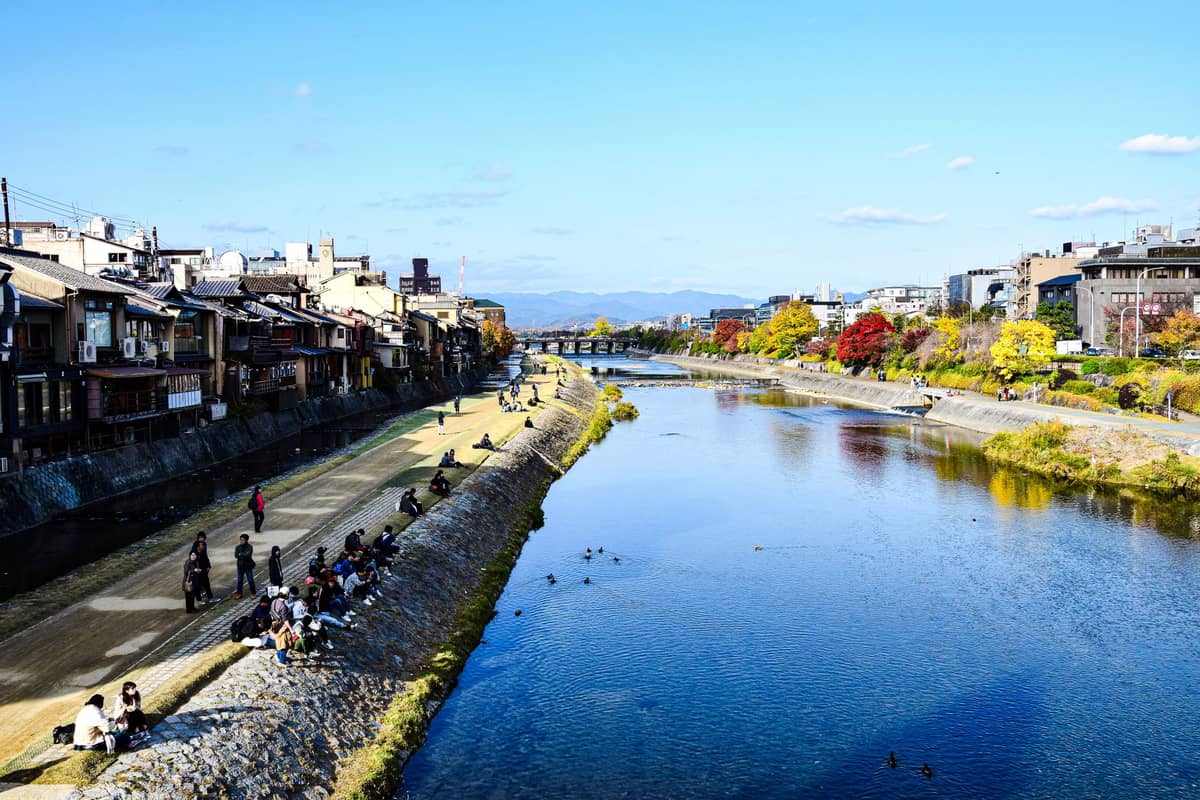 A wide view of the Kamogawa River in Kyoto with traditional and modern buildings along the riverside and people relaxing on both banks.