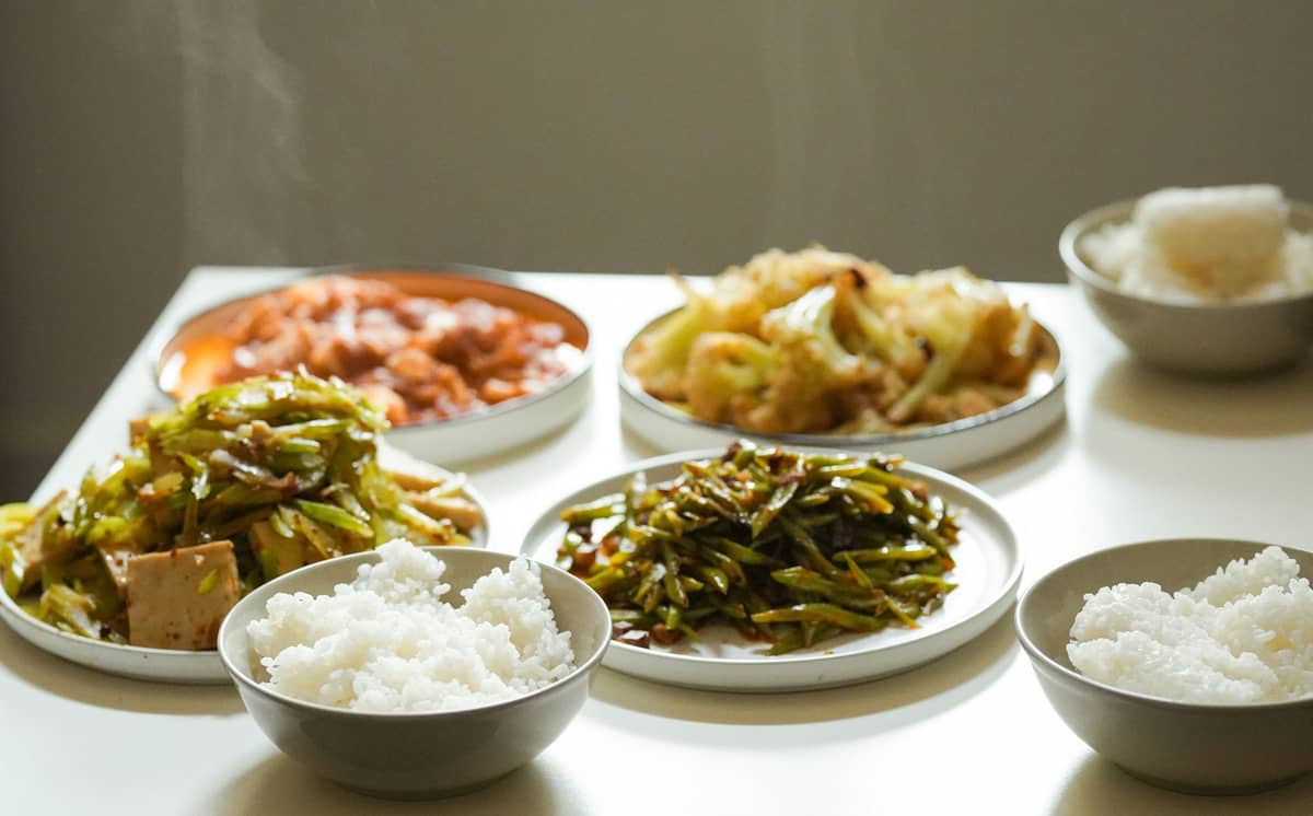 A selection of small home-cooked Japanese dishes including rice, greens, tofu, and pickles on a table.