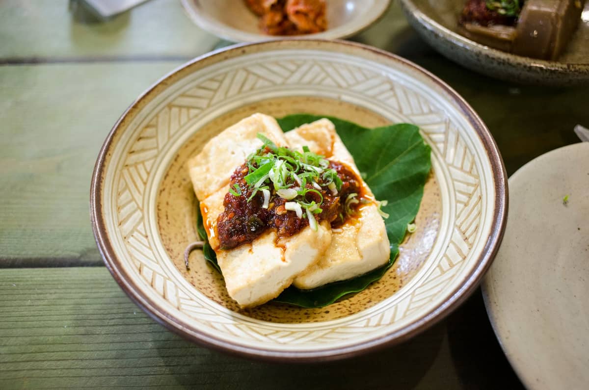A shallow bowl with silken tofu topped with a savory sauce and chopped green onions, served on a green leaf at a wooden table.