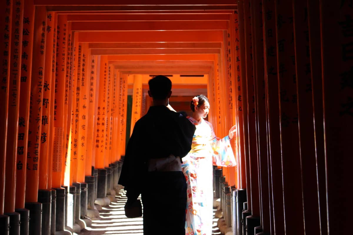 A couple in kimono stands under the famous red torii gates of Fushimi Inari shrine in Kyoto, bathed in sunlight.