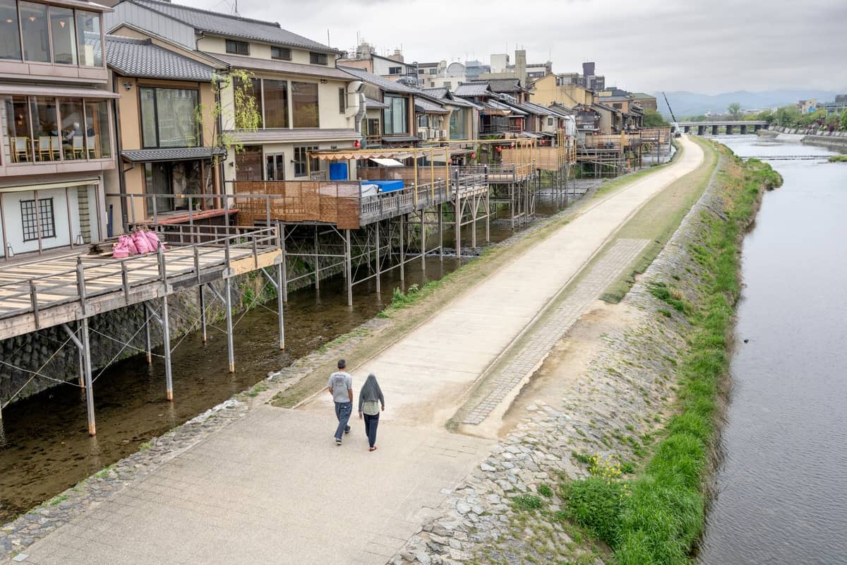 Couple walking along the Kamogawa River in Kyoto beside traditional wooden buildings with decks overhanging the water on a cloudy day.