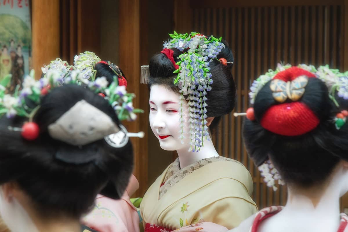 A group of maiko in traditional kimono and elaborate hair ornaments gathered outdoors in Kyoto, with one maiko facing forward and others seen from behind.
