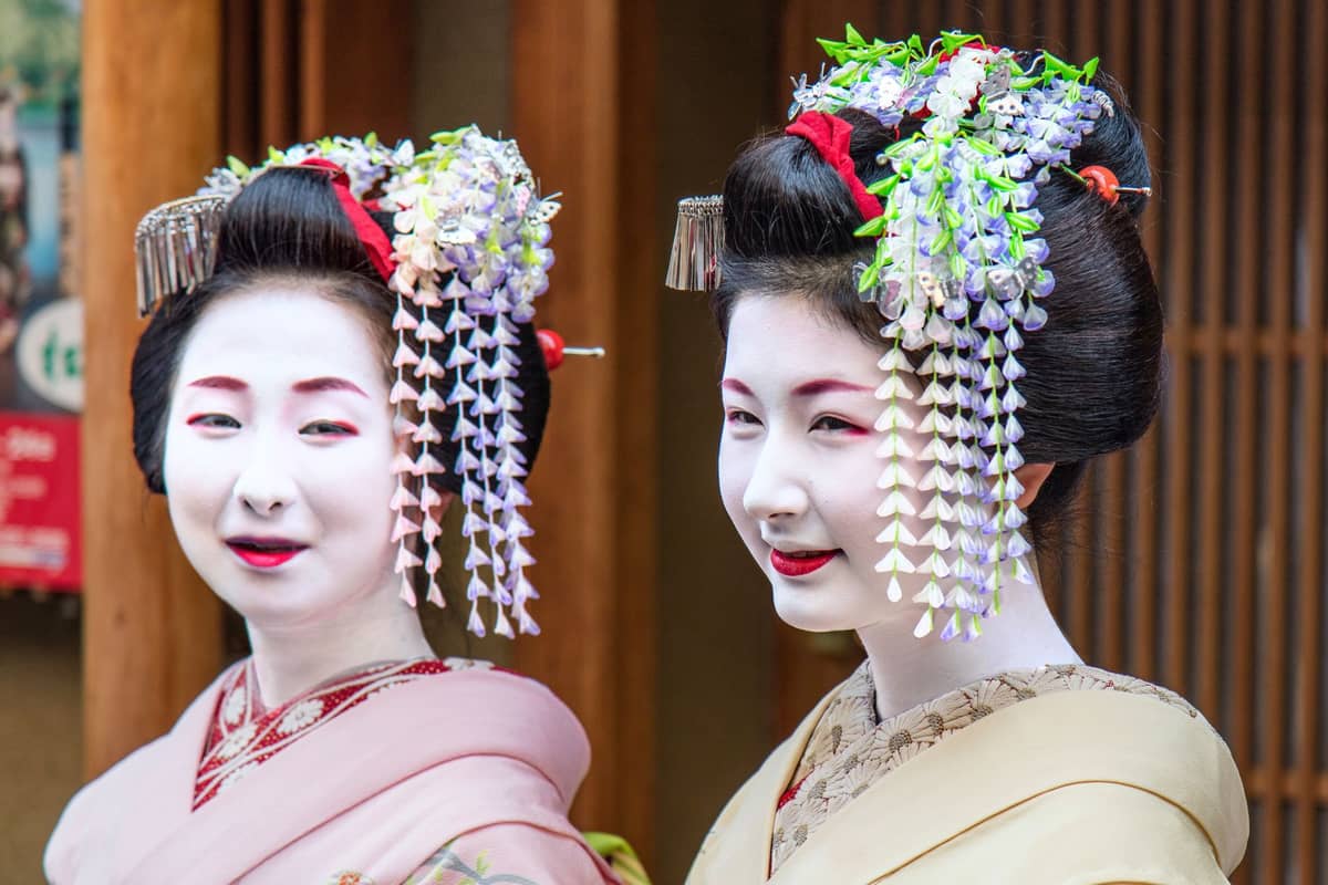 Two women dressed as maiko in traditional kimono, with elaborate hairpieces and white face makeup, standing in front of wooden slats in Kyoto.