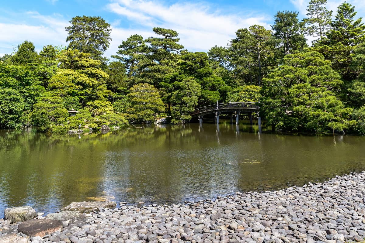 A serene pond with a traditional stone bridge surrounded by lush green trees inside Kyoto Gyoen National Garden on a sunny day.