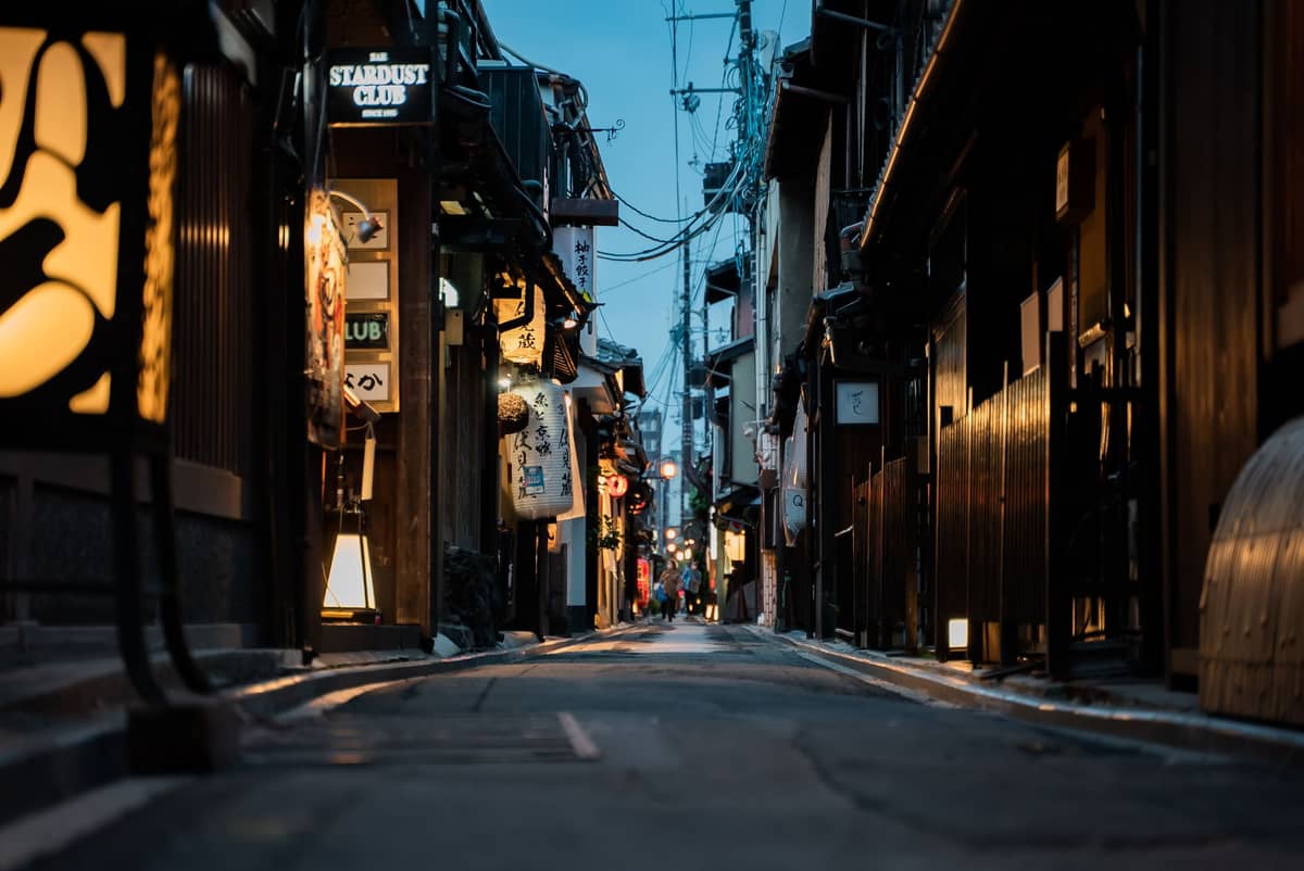 Narrow atmospheric lane in Kyoto at dusk, lined with traditional wooden buildings, lanterns, and subtle shop signs, inviting nighttime exploration.