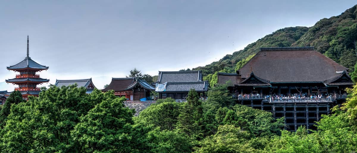 Panoramic view of Kiyomizu-dera temple complex surrounded by lush green forested mountains under a cloudy sky in Kyoto.