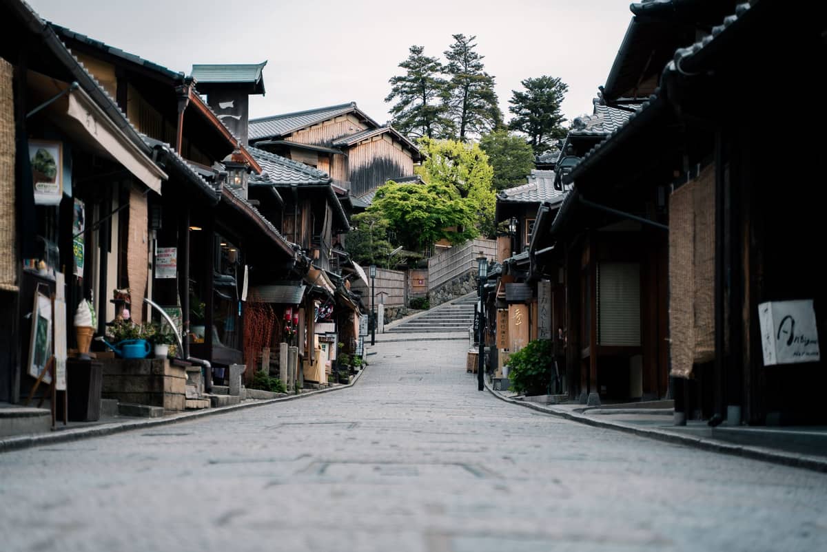 Quiet atmospheric alleyway in Kyoto’s historic district with traditional wooden buildings lining a gently sloping stone street and lush greenery in the background.