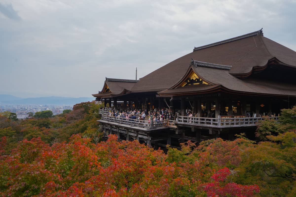 Large crowd on the main Kiyomizu-dera temple balcony surrounded by autumn foliage in Kyoto, with city and mountains in the background.
