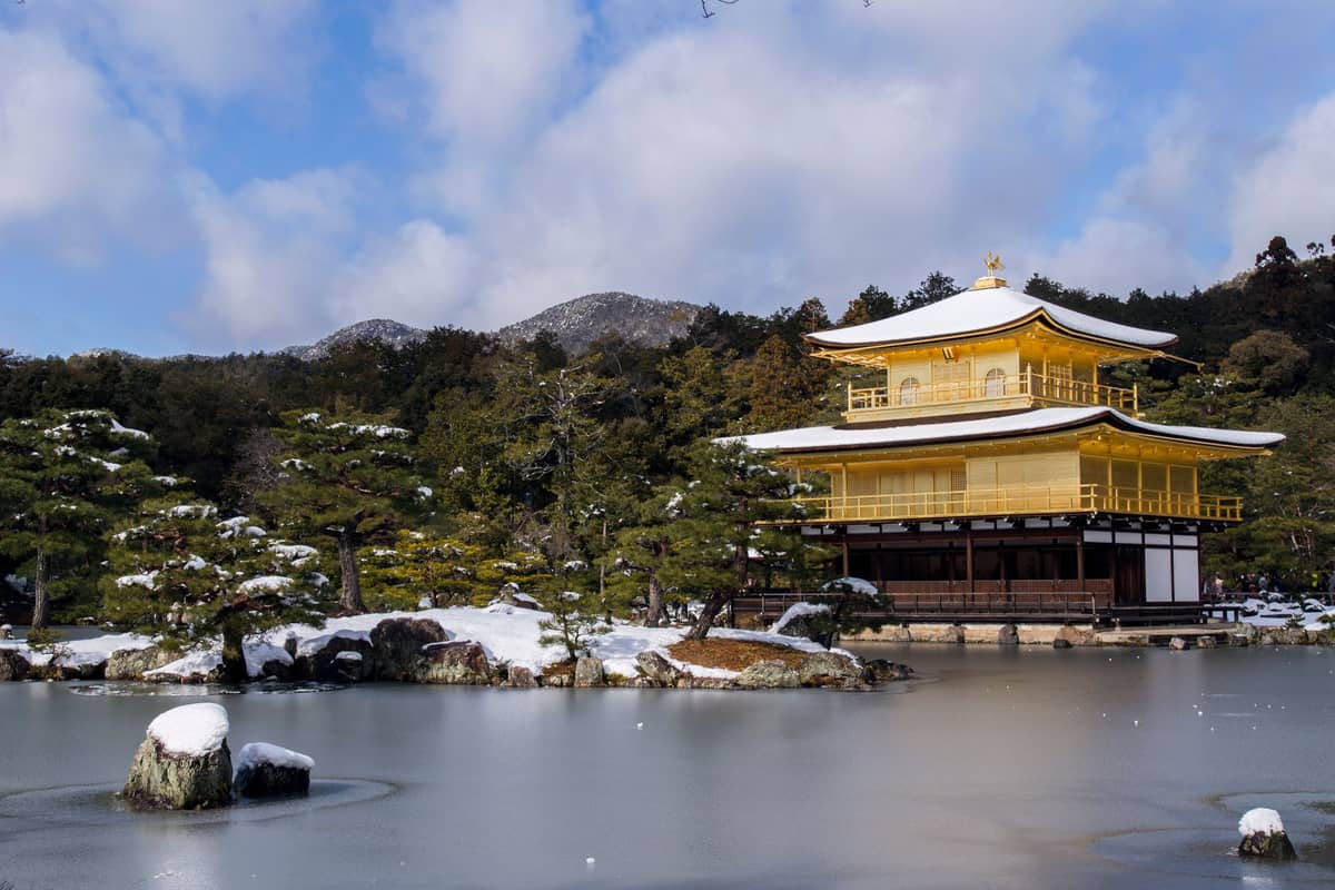 Kinkaku-ji, Kyoto’s famous Golden Pavilion, reflected in a snowy pond landscape under a clear winter sky, surrounded by pine trees and distant mountains.