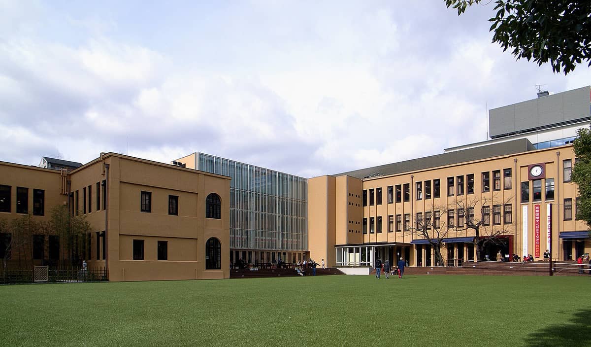 Wide view of the Kyoto International Manga Museum's exterior, showing its distinct tan facade, modern glass extension, and open green lawn in front.