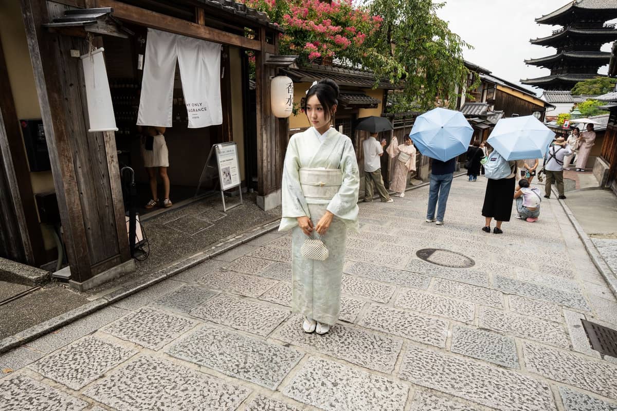 Woman in kimono stands on a historic stone street in Kyoto with traditional wooden buildings and a five-story pagoda in the background.