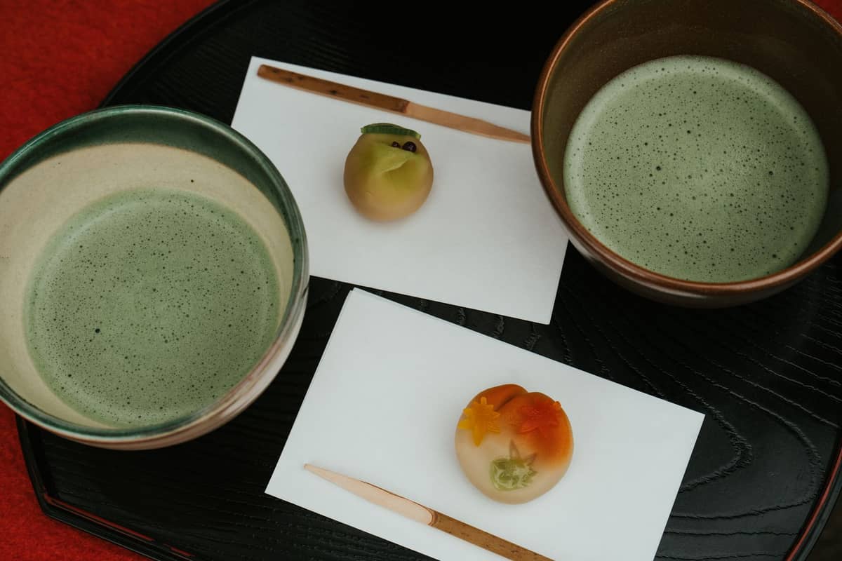 Overhead view of two bowls of matcha tea with traditional Japanese wagashi sweets on paper, set on a black lacquer tray. This represents Kyoto matcha culture.