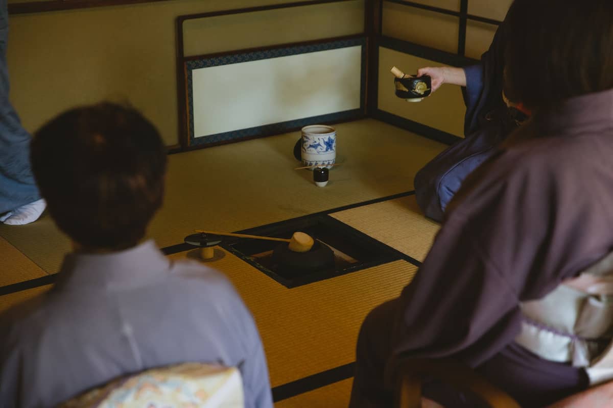 People dressed in kimono participate in a traditional Japanese tea ceremony in a tatami room, with tea utensils arranged on the floor.
