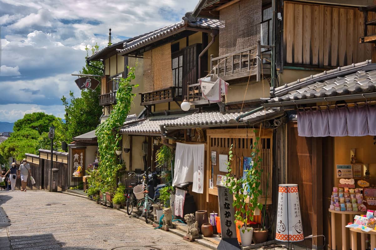 Traditional wooden buildings along a narrow stone street in Kyoto, with signs and shopfronts, on a sunny day.