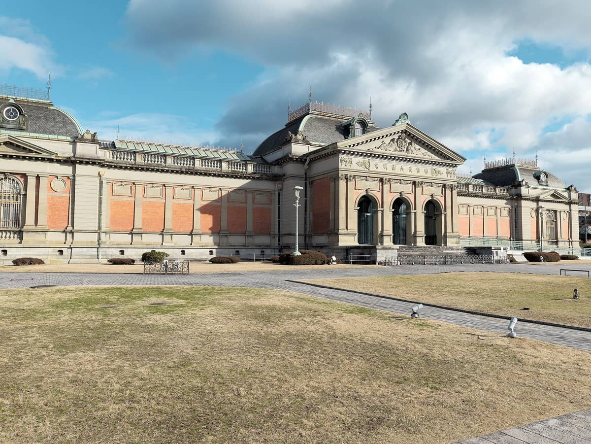 Wide landscape view of the Meiji-era main building of the Kyoto National Museum under blue sky, with green lawn and decorative architectural details in front.