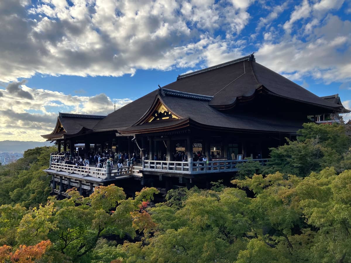 Wide view of Kiyomizudera Temple's main hall perched above a forest of green trees in Kyoto under a partly cloudy sky.